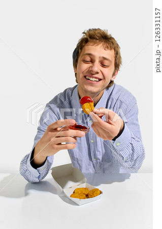 Smiling young man dipping crispy nugget into sauce with cheerful expression, holding box of nuggets on table against white background. Smiling young man dipping crispy nugget into sauce with cheerful expression, holding box of nuggets on table against white background. 126331157