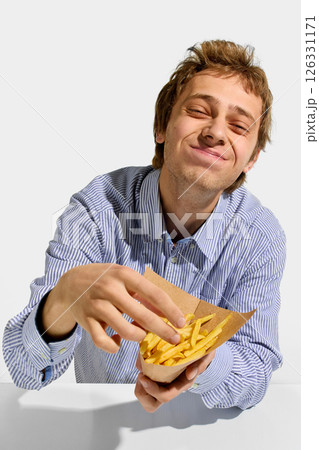 Man smiling with full cheeks while grabbing crispy French fries from paper cone against white background. Joyful food moment. Man smiling with full cheeks while grabbing crispy French fries from paper cone against white background. Joyful food moment. 126331171