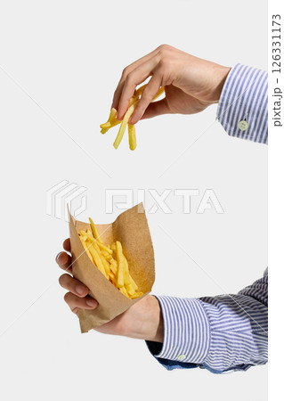 One hand picking up crispy French fries while other holds full brown paper cone, striped shirt visible against white background. 126331173