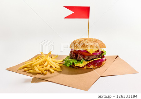 Juicy cheeseburger with lettuce, tomato, onions, ketchup, and cheddar topped with red flag stands next to crispy french fries on brown parchment paper. Juicy cheeseburger with lettuce, tomato, onions, ketchup, and cheddar topped with red flag stands next to crispy french fries on brown parchment paper. 126331194