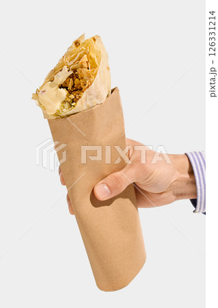 Shawarma roll with grilled meat, white sauce, cabbage, and flatbread held in brown paper sleeve, viewed from hand against white background. Shawarma roll with grilled meat, white sauce, cabbage, and flatbread held in brown paper sleeve, viewed from hand against white background. 126331214