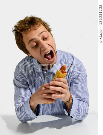 Man in blue striped shirt standing with one hand holding sausage roll and looking up joyfully before bite with playful wide-open mouth expression. Man in blue striped shirt standing with one hand holding sausage roll and looking up joyfully before bite with playful wide-open mouth expression. 126331232