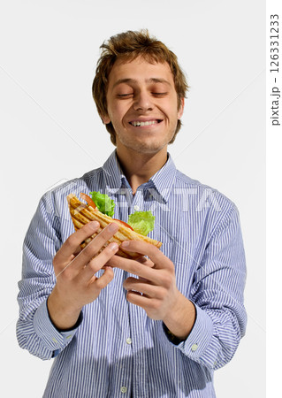 Man smiling with closed eyes while gently holding grilled sandwich filled with lettuce and tomato, experiencing pleasure from taste against white background 126331233