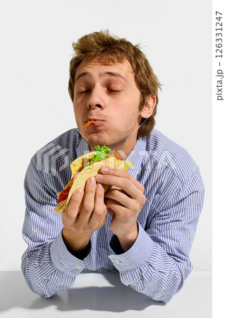 Man chewing flavorful taco with eyes closed, lettuce and carrot spilling from tortilla in happy food moment against white background. 126331247