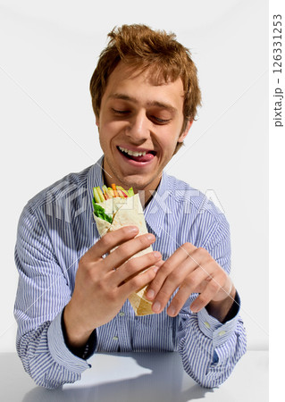 Happy young man, in striped shirt, holding vegetable wrap with playful tongue out, enjoying colorful healthy snack against white background. Happy young man, in striped shirt, holding vegetable wrap with playful tongue out, enjoying colorful healthy snack against white background. 126331253
