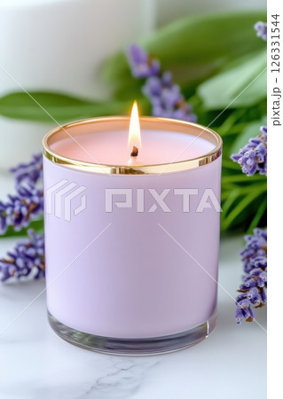 An elegant lavender-colored scented candle in a luxurious glass vessel with a gold rim, captured against a white marble background. The lit candle emits a warm glow. 126331544