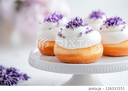 Delicate mini donuts topped with smooth white frosting and garnished with fresh lavender buds, presented on an elegant white ceramic cake stand.  Delicate mini donuts topped with smooth white frosting and garnished with fresh lavender buds, presented on an elegant white ceramic cake stand.  126331551
