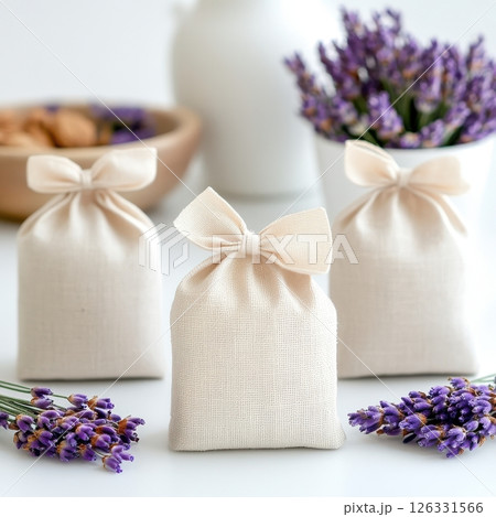 Elegant composition featuring cream-colored linen sachets with delicate bow ties, accompanied by fresh lavender sprigs. The image showcases three fabric pouches against a clean white background. 126331566