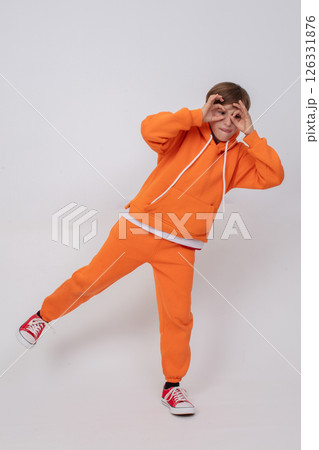Boy in orange sweatshirt and orange pants making glasses sign with hands. Boy standing on grey background Boy in orange sweatshirt and orange pants making glasses sign with hands. Boy standing on grey background 126331876