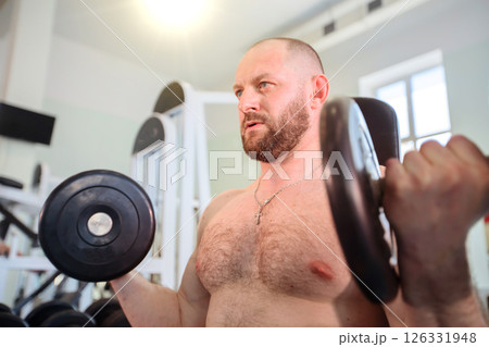 A male athlete lifts weights in the gym. He is bald and has a beard. 126331948