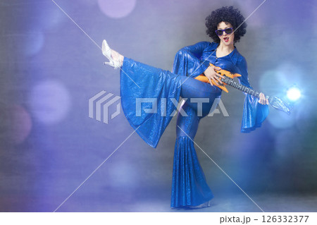 Fun eighties style. A girl posing in a black wig with a toy guitar on a gray background. Fun eighties style. A girl posing in a black wig with a toy guitar on a gray background. 126332377