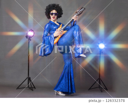 Fun eighties style. A girl posing in a black wig with a toy guitar on a gray background. Fun eighties style. A girl posing in a black wig with a toy guitar on a gray background. 126332378