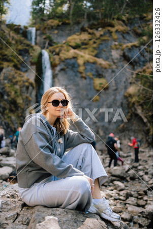 Blonde woman wearing sunglasses and casual clothing sitting on rock near waterfall in mountains, enjoying beautiful scenery and fresh air. Blonde woman on mountainside rock with a waterfall behind Blonde woman wearing sunglasses and casual clothing sitting on rock near waterfall in mountains, enjoying beautiful scenery and fresh air. Blonde woman on mountainside rock with a waterfall behind 126332426