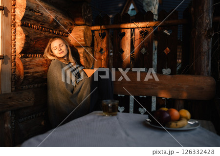Cozy atmosphere in wooden mountain chalet with young woman relaxing, wrapped in blanket, enjoying sunset light and plate of fresh fruit with cup of tea. Young woman basks in warm rays of sunset Cozy atmosphere in wooden mountain chalet with young woman relaxing, wrapped in blanket, enjoying sunset light and plate of fresh fruit with cup of tea. Young woman basks in warm rays of sunset 126332428