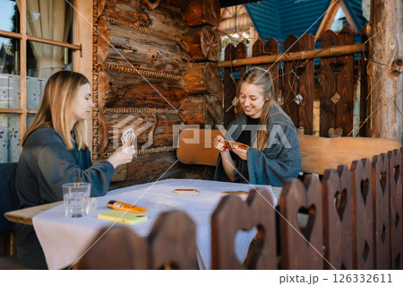 Two friends sitting at rustic wooden table on porch of cozy mountain chalet, playing cards and savoring their leisure time together in beautiful outdoors. Friends enjoying card game on mountain chalet 126332611