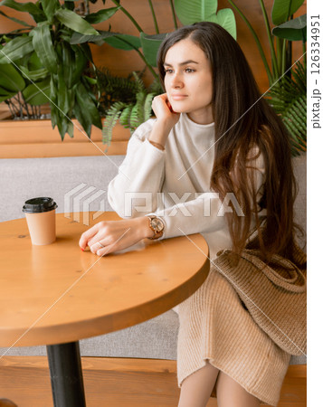 Elegant woman with coffee sitting in cafe, surrounded by green plants, wearing neutral toned stylish outfit Elegant woman with coffee sitting in cafe, surrounded by green plants, wearing neutral toned stylish outfit 126334951