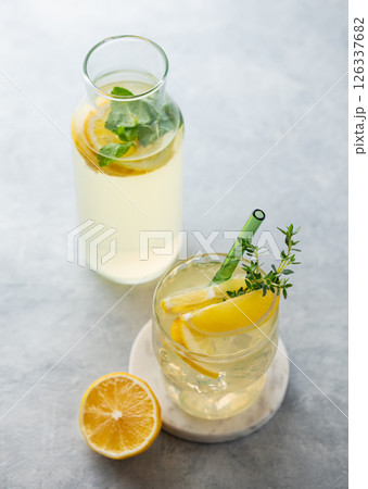 Lemonade with fresh lemon, thyme and ice on a light background with bottle and sunlight. Lemonade with fresh lemon, thyme and ice on a light background with bottle and sunlight. 126337682