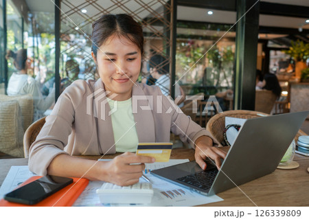 Woman using a laptop and credit card for online shopping in a cozy cafe, surrounded by documents and tech devices. 126339809