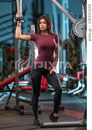 fit woman stands confidently by a cable machine in a gym showcasing her commitment to strength training while wearing athletic attire. The environment is well equipped and modern. 126341829