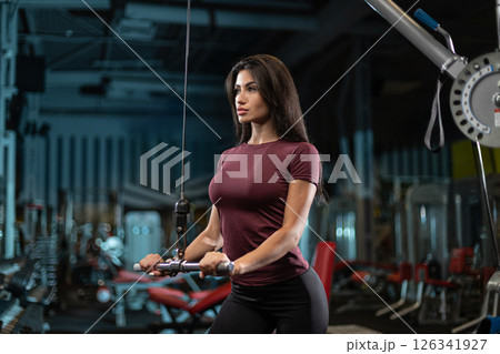 woman is focused on her workout as she uses a cable machine in a well equipped gym. The environment is spacious with various fitness equipment visible in the background. woman is focused on her workout as she uses a cable machine in a well equipped gym. The environment is spacious with various fitness equipment visible in the background. 126341927