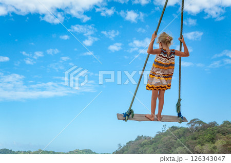 Happy girl have fun swinging high in mid air. Flying up upside down on rope swing on sea beach. Travel adventure on paradise tropical island. Family lifestyle, activity on summer vacation with kids. Happy girl have fun swinging high in mid air. Flying up upside down on rope swing on sea beach. Travel adventure on paradise tropical island. Family lifestyle, activity on summer vacation with kids. 126343047