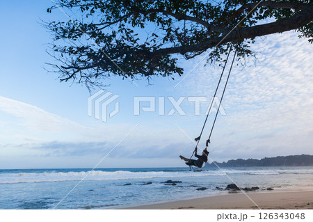 Happy girl have fun swinging high in mid air. Flying on rope swing on sea beach. Travel adventure on paradise tropical island. Family lifestyle, activity on summer vacation. Happy girl have fun swinging high in mid air. Flying on rope swing on sea beach. Travel adventure on paradise tropical island. Family lifestyle, activity on summer vacation. 126343048