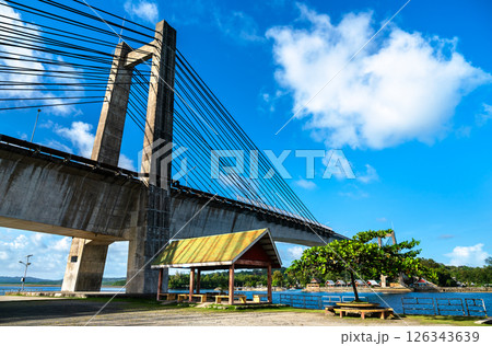 Koror - Babeldaob Japan - Palau Friendship Bridge under a bright blue sky. The modern cable-stayed structure spans a calm turquoise channel, with a shaded seating area, coastal trees and traditional 126343639