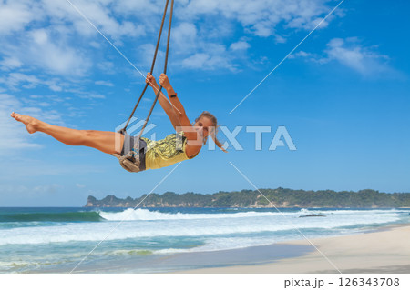 Happy girl have fun swinging high in mid air. Flying on rope swing on sea beach. Travel adventure on paradise tropical island. Family lifestyle, activity on summer vacation. 126343708