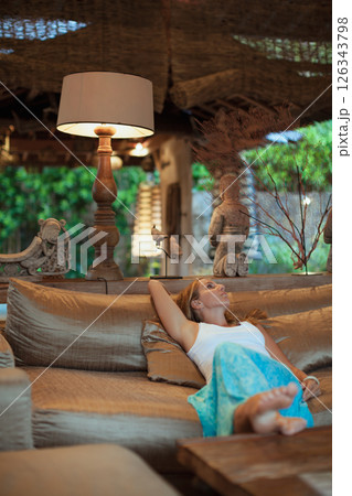 Young woman sit on sofa inside luxury wooden villa. Girl relaxing on family summer vacation. Tropical lifestyle. 126343798