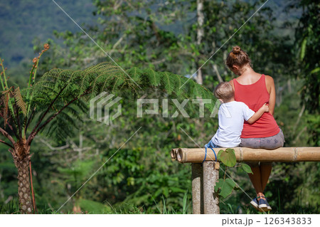 Mother, little baby son have fun outdoors, sit on bench under the tree on sunny lawn. Happy family lifestyle. Pretty young mom hugging her child in park outside. Summer camp walks on vacation. 126343833