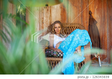 Young woman sit on open-air veranda of wooden bungalow with tropical garden view. Girl relaxing in luxury villa on family summer vacation. Countryside lifestyle. 126343839