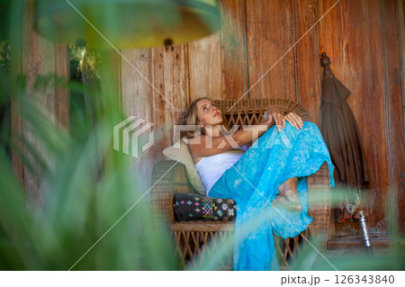 Young woman sit on open-air veranda of wooden bungalow with tropical garden view. Girl relaxing in luxury villa on family summer vacation. Countryside lifestyle. 126343840