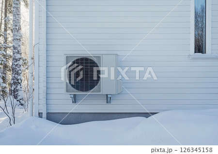 Outdoor heat pump covered in snow near a cozy house, illustrating winter home heating and energy efficiency. 126345158