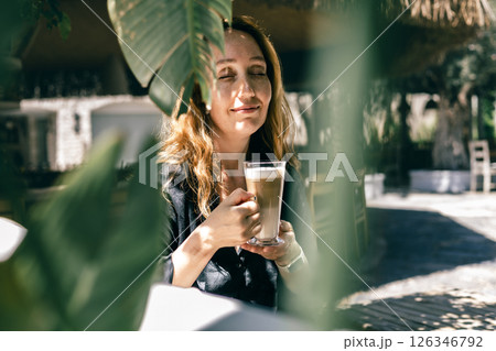 Peaceful Woman Enjoying Latte in Tropical Café Through Green Leaves on a Sunny Morning 126346792
