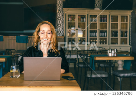 Smiling woman using laptop in a trendy cafe, enjoying remote work with a drink. Cozy workspace  126346796