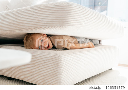 Child playfully hides between the cushions of a white sofa, displaying a curious and mischievous expression. The cozy setting suggests a fun and imaginative moment of play. Child playfully hides between the cushions of a white sofa, displaying a curious and mischievous expression. The cozy setting suggests a fun and imaginative moment of play. 126351579