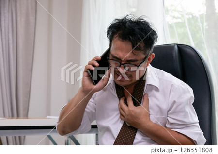 Frustrated businessman gripping chest in anxiety while talking on phone, stressed expression with wrinkled face, wearing glasses and loosened tie, sitting indoors in office Frustrated businessman gripping chest in anxiety while talking on phone, stressed expression with wrinkled face, wearing glasses and loosened tie, sitting indoors in office 126351808