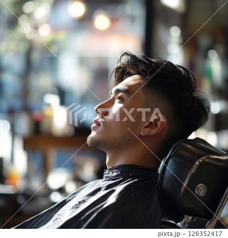 Asian male is seated in barber shop chair, eyes closed, enjoying haircut, surrounded by stylish decor and warm lighting, creating a tranquil environment 126352417