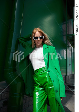 High fashion photo of a young blond woman in green coat and leather pants. Monochrome green outfit, white glasses and shoes. High fashion photo of a young blond woman in green coat and leather pants. Monochrome green outfit, white glasses and shoes. 126353017