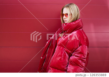 High fashion photo of a young blond woman in red glossy down jacket, poloneck, and pants. Monochrome trendy red outfit. 126353869