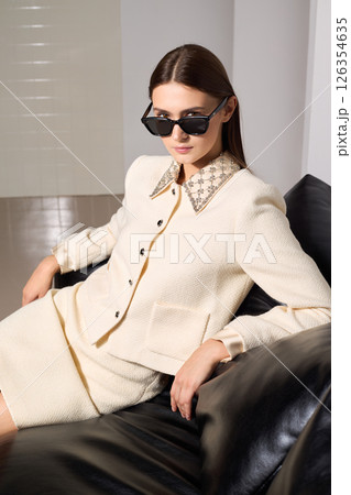 Stylish female fashion model sits on sofa confidently in a trendy beige outfit and sunglasses indoors 126354635