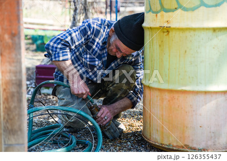 Elderly Russian man sitting near a barrel pulling a drainage hose to the side with visible tension and effort in a quiet rural setting. Elderly Russian man sitting near a barrel pulling a drainage hose to the side with visible tension and effort in a quiet rural setting. 126355437
