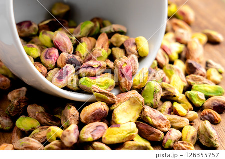 Peeled pistachio kernels in white cup are placed in pile of natural nutritious nuts. Macro shot Peeled pistachio kernels in white cup are placed in pile of natural nutritious nuts. Macro shot 126355757