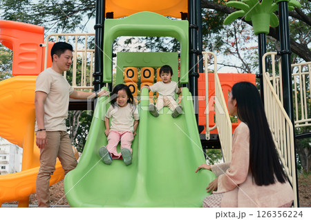 Smiling siblings enjoy a fun ride down a double slide as their parents stand by 126356224