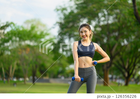 Portrait of a cheerful female athlete lifting dumbbells in a green park 126356226