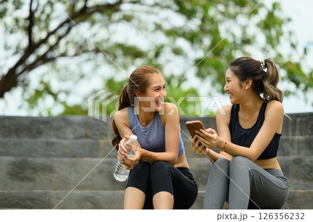Sporty women relax on stone steps, sharing a joyful moment after a workout Sporty women relax on stone steps, sharing a joyful moment after a workout 126356232