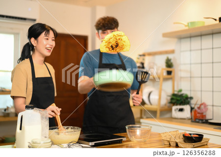 Playful couple flipping pancake, sharing laughter during home cooking 126356288