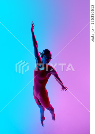 Female swimmer in red suit performs elegant leap in vibrant pink and blue light studio, suspended midair with stretched arm, reflecting power and finesse. 126356612