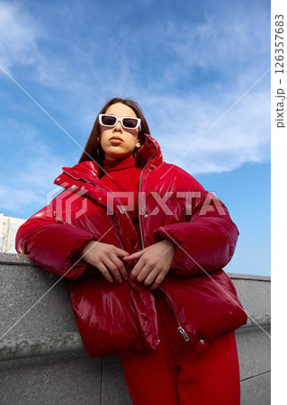 High fashion photo of a young brunette woman in red glossy down jacket, and red pants. Urban street fashion. High fashion photo of a young brunette woman in red glossy down jacket, and red pants. Urban street fashion. 126357683