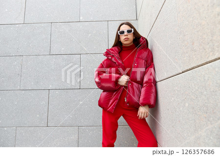 High fashion photo of a young brunette woman in red glossy down jacket, and red pants. Urban street fashion. High fashion photo of a young brunette woman in red glossy down jacket, and red pants. Urban street fashion. 126357686